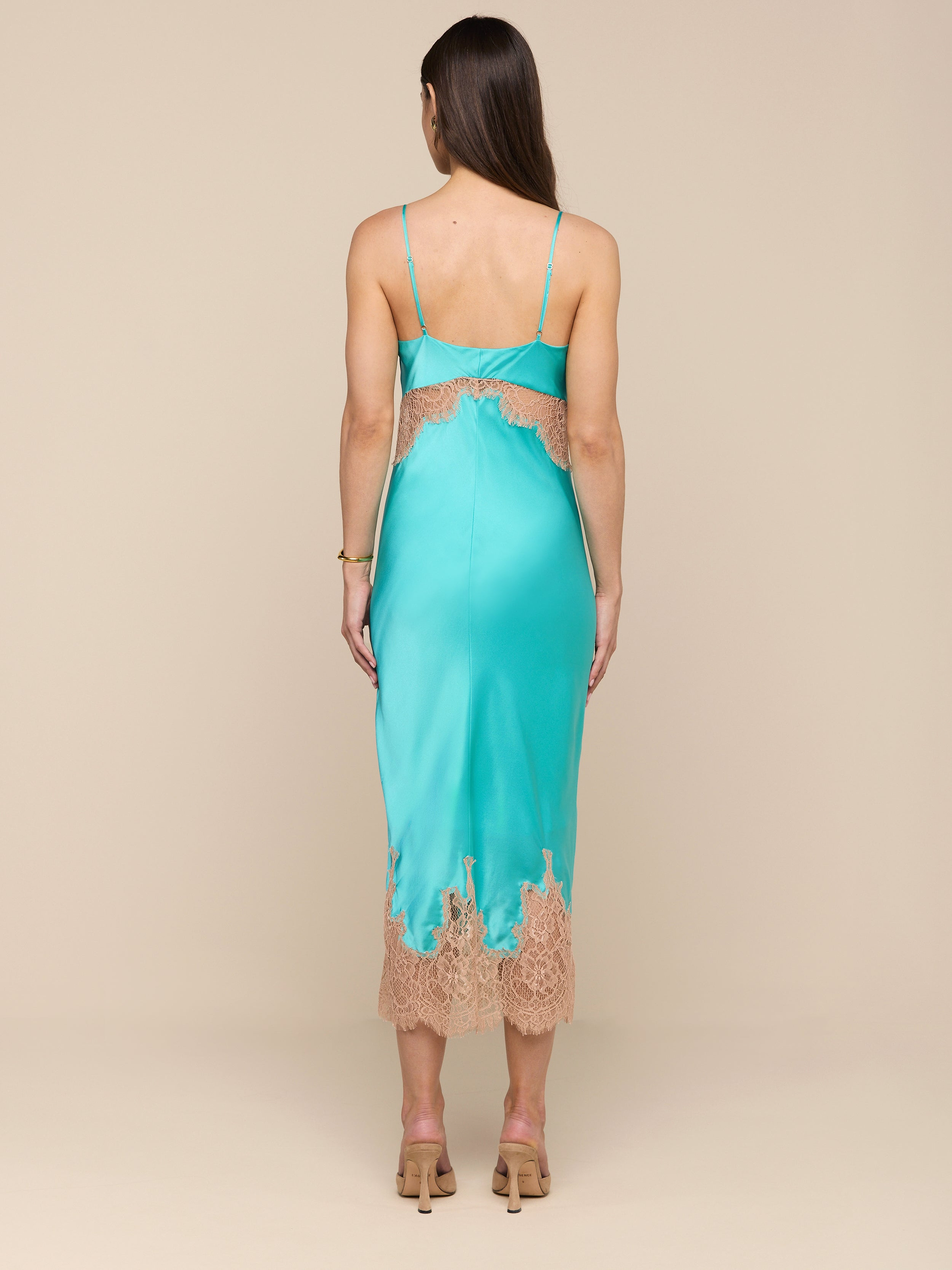 A woman stands facing away against a plain white background, wearing the Scotly Silk Lace Dress—a bias-cut teal silk midi dress featuring lace insets at the hem and back—paired with beige heeled sandals.