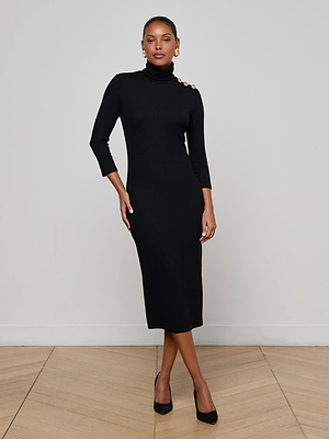 A woman wears the L’AGENCE Voletta Turtleneck Knit Dress—fitted, black, long-sleeve with gold shoulder buttons—in soft microfiber modal jersey. She stands on a light wood floor against a white wall, pairing it with black heels for fall to winter.