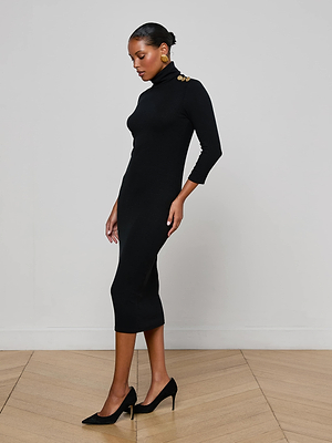 A woman in the L'AGENCE Voletta Turtleneck Knit Dress, a fitted black essential in soft microfiber modal jersey, stands in profile on a wooden floor, paired with black heels and gold earrings against a plain light background.