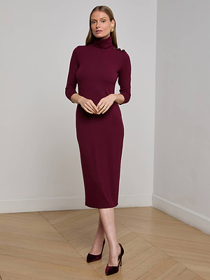 A woman indoors wears the L'AGENCE Voletta Turtleneck Midi Dress—a fitted, maroon stretch modal midi with button-shoulder detail—paired with burgundy heels and gold earrings on a light wood floor.