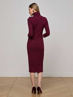 A woman with straight, light brown hair stands on a wooden floor facing away, wearing the L'AGENCE Voletta Turtleneck Midi Dress in dark maroon and coordinating velvet heels, set against a plain wall.