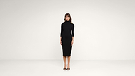 A woman stands facing forward in a plain white room, wearing a black, form-fitting, knee-length dress with three-quarter sleeves and black high heels.