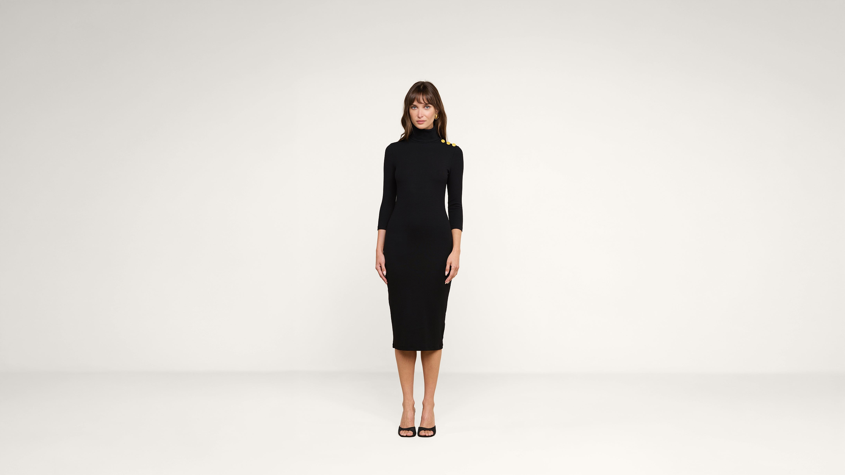 A woman stands facing forward in a plain white room, wearing a black, form-fitting, knee-length dress with three-quarter sleeves and black high heels.