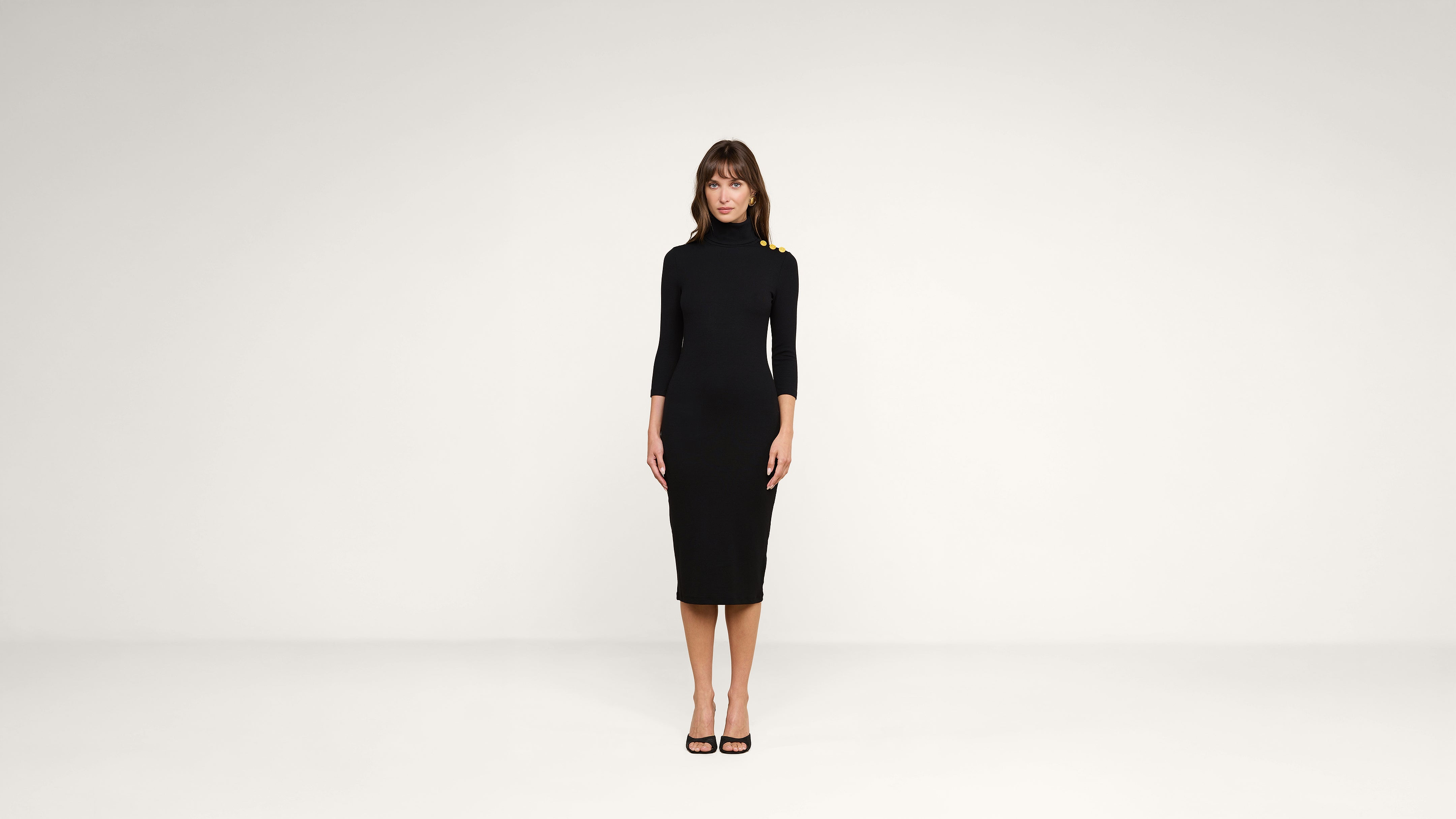 A woman stands facing forward in a plain white room, wearing a black, form-fitting, knee-length dress with three-quarter sleeves and black high heels.