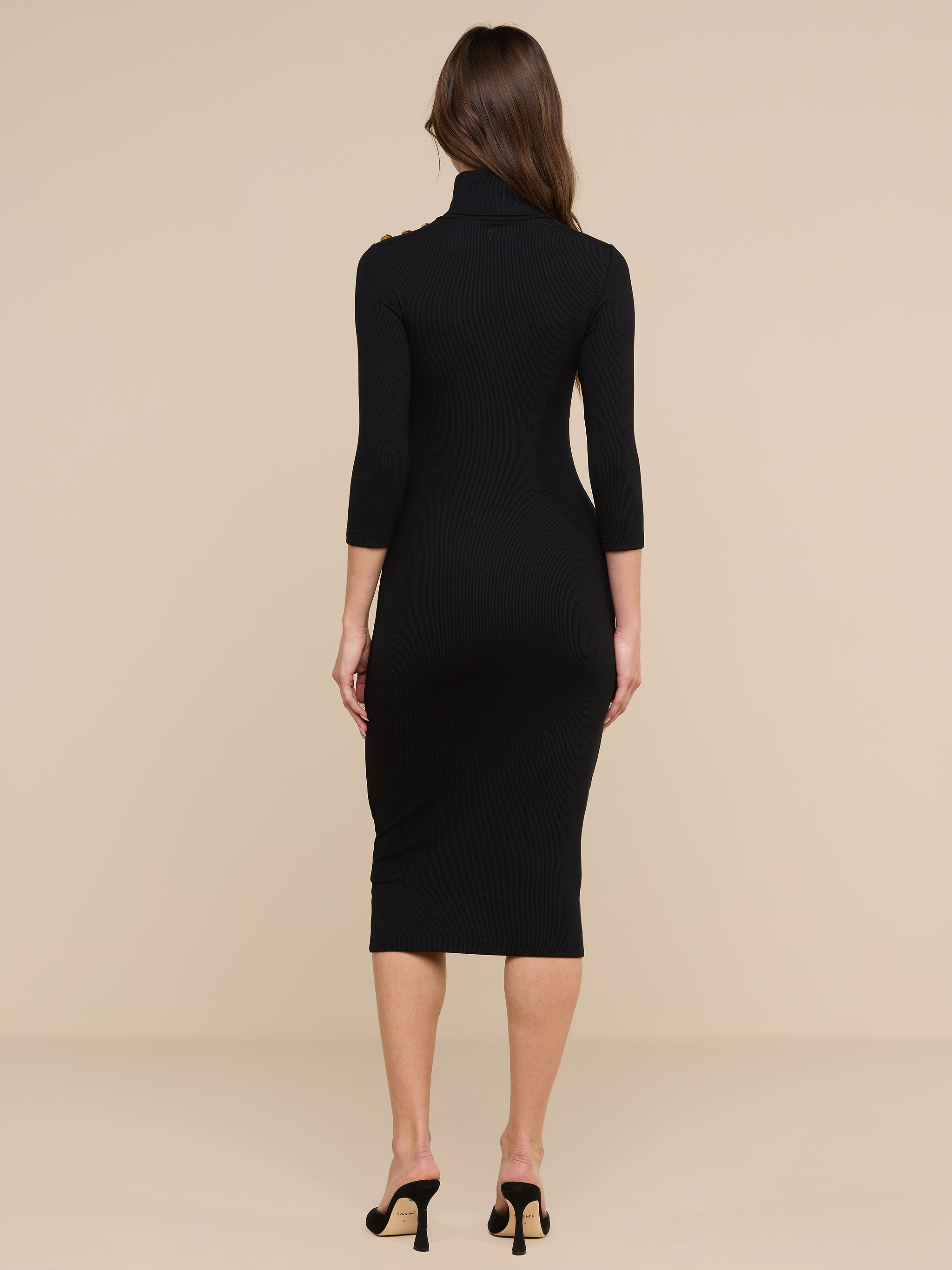 Une femme aux longs cheveux châtains se tient dos à la caméra, vêtue de la robe en maille à col roulé Voletta et de talons noirs à bout ouvert — un incontournable ajusté et polyvalent de la saison automne-hiver —, sur un fond beige.
