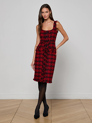 A woman stands against a plain wall wearing the L'AGENCE Aya Tweed Midi Dress in red and black plaid, styled with black tights and heels. Her long brown hair and neutral expression complete this chic Fall 25 NYFW look.