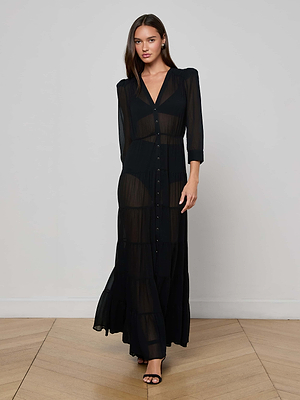 A woman with long, straight hair stands indoors on a wooden floor, facing forward and wearing the L'AGENCE Seraphina Tiered Maxi Dress with buttons down the front, long sleeves, a deep V-neckline, and black heeled sandals.