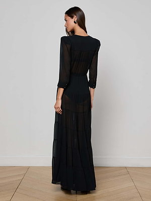 A woman with straight, loose hair and earrings stands on a wooden floor facing away, wearing the sheer black Seraphina Tiered Maxi Dress by L'AGENCE. The background is a plain, light-colored wall.