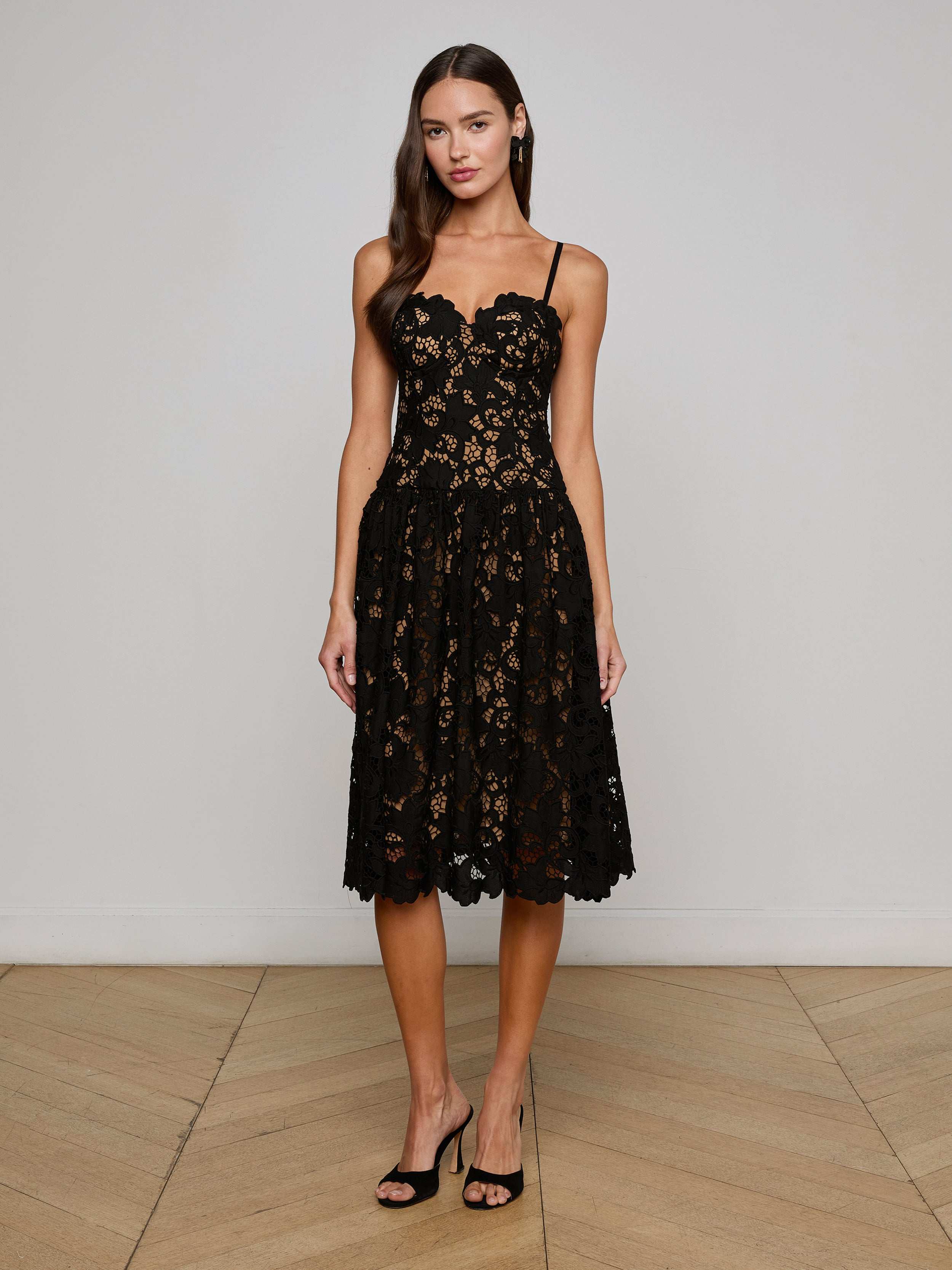 A woman with long, wavy brown hair poses indoors against a light wall and wooden floor, wearing the L'AGENCE Sale Flyn Lace Midi Dress— a sleeveless black floral eyelet lace dress with a scalloped neckline—paired with black heels.