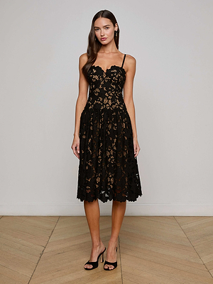 A woman with long, wavy brown hair poses indoors against a light wall and wooden floor, wearing the L'AGENCE Sale Flyn Lace Midi Dress— a sleeveless black floral eyelet lace dress with a scalloped neckline—paired with black heels.
