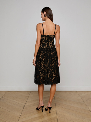 A woman faces away from the camera in the L'AGENCE Sale Flyn Lace Midi Dress, a black fit-and-flare design with thin straps and delicate floral eyelet lace, paired with high heels. She stands before a plain light wall and wooden floor.