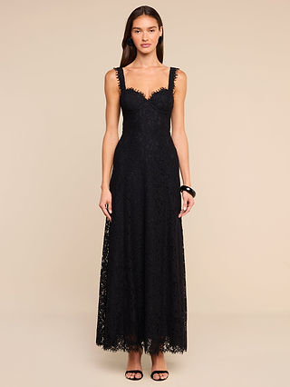 A woman stands against a plain beige background wearing the Sutton Lace Maxi Dress—a sleeveless, floor-length black lace gown with a sweetheart neckline, underwire cups, and scalloped edges. She has long hair styled down and accessorizes with black strappy heels and a black bracelet.