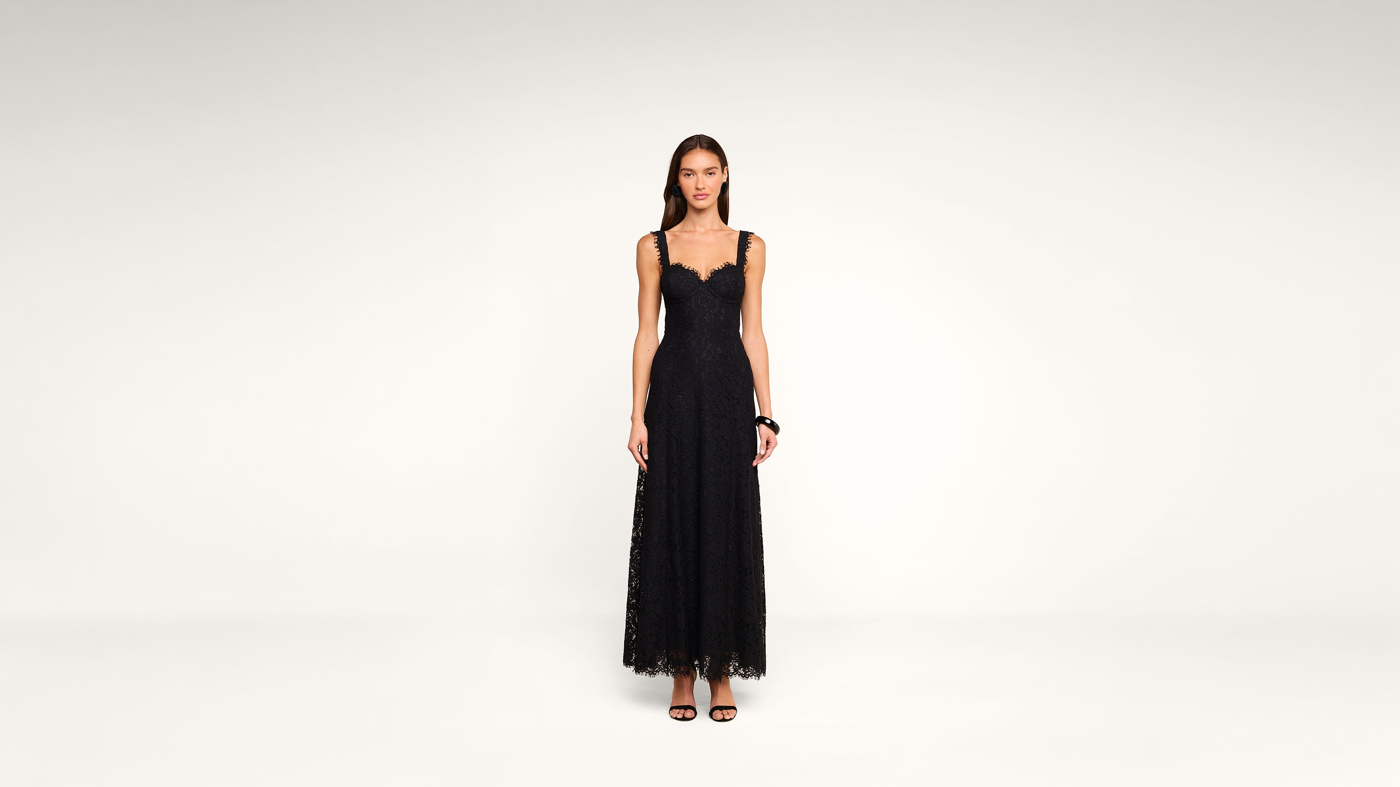Woman standing against a plain background, wearing a long, sleeveless black lace dress with a scalloped neckline and black high-heeled sandals.