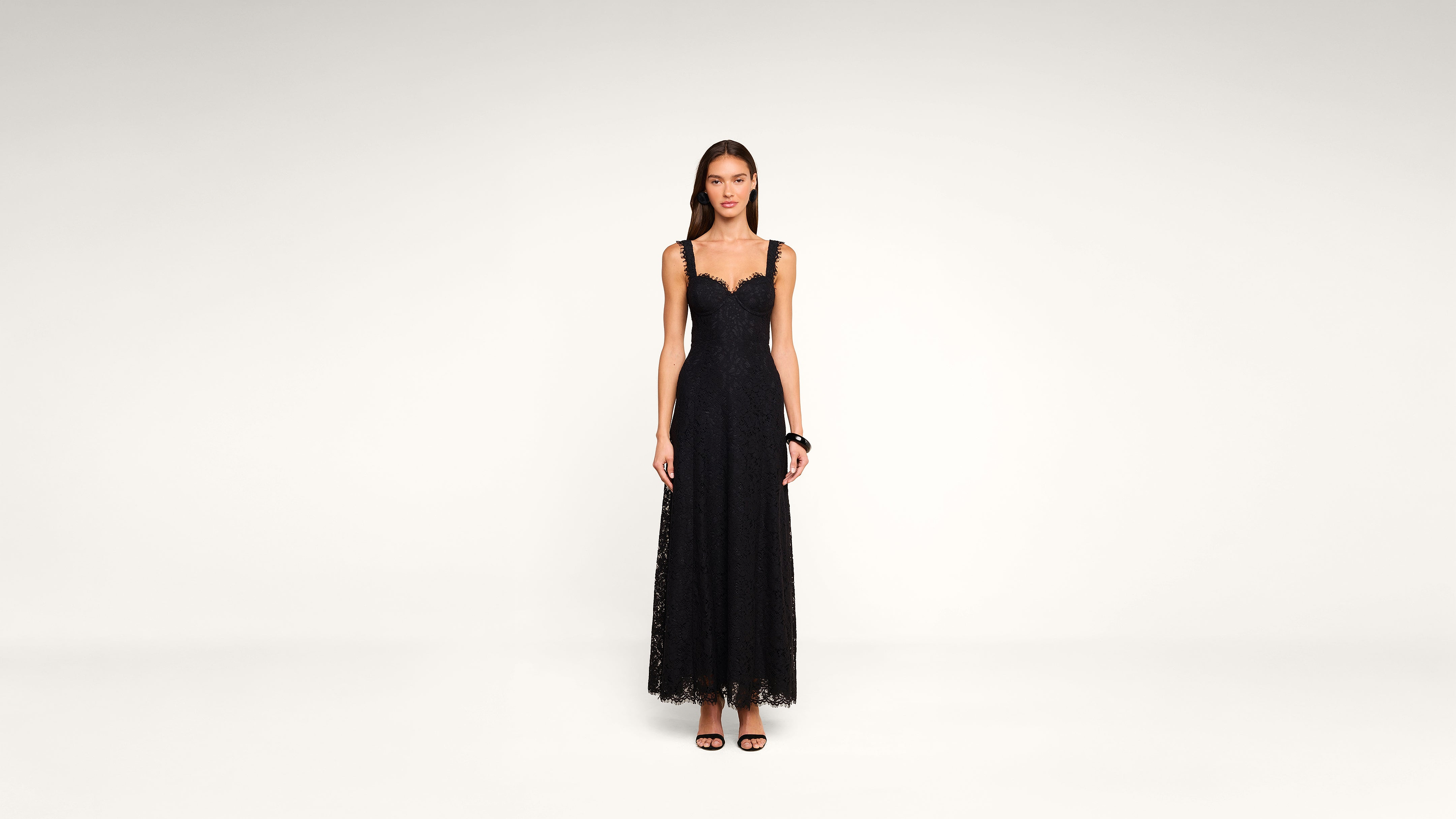 Woman standing against a plain background, wearing a long, sleeveless black lace dress with a scalloped neckline and black high-heeled sandals.