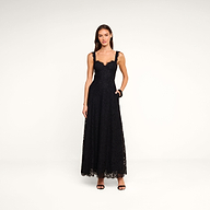 A woman stands against a plain white background, wearing the Sutton Lace Maxi Dress—an elegant black lace maxi dress with thin straps—paired with black high-heeled sandals. She has long brown hair, and her left hand rests in her dress pocket.