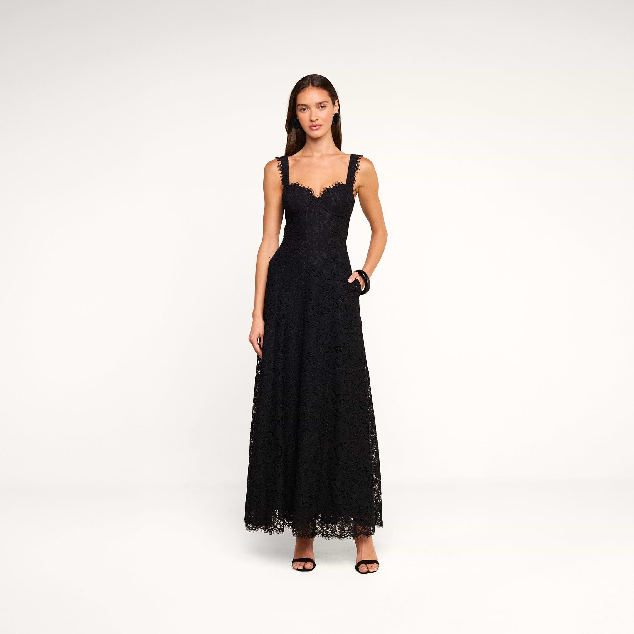 A woman stands against a plain white background, wearing the Sutton Lace Maxi Dress—an elegant black lace maxi dress with thin straps—paired with black high-heeled sandals. She has long brown hair, and her left hand rests in her dress pocket.
