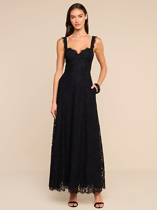 A woman poses against a beige background, showcasing the Sutton Lace Maxi Dress—a sleeveless black lace gown with underwire cups, a sweetheart neckline, and scalloped edges. The floor-length floral lace design highlights her long brown hair and pairs elegantly with her black open-toe heels.