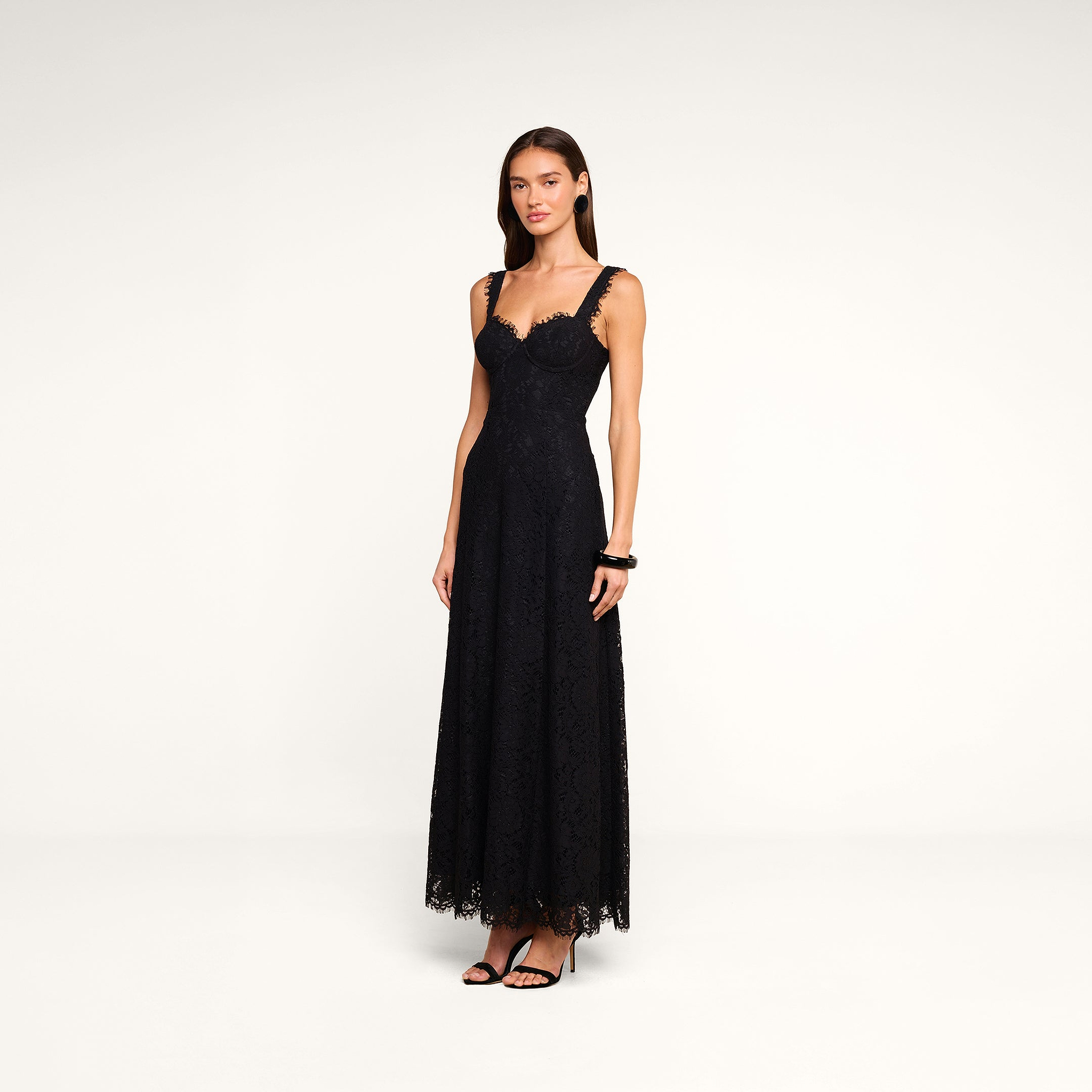 A woman stands on a plain background wearing the Sutton Lace Maxi Dress with thin straps and black heels. She has straight, dark hair and accessorizes with bracelets.