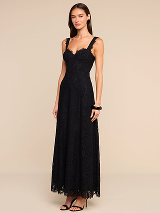 A woman stands against a plain beige background wearing the Sutton Lace Maxi Dress—a sleeveless, floor-length black lace gown with a fitted bodice featuring underwire cups and a flared skirt—accessorized with black bracelets and high-heeled sandals.