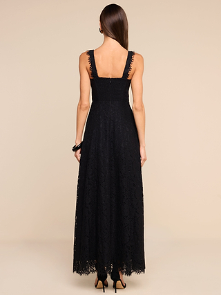 A woman with straight brown hair stands facing away, wearing the Sutton Lace Maxi Dress—a long black lace maxi dress featuring wide straps and a square neckline. She completes her look with black heels, set against a beige background.