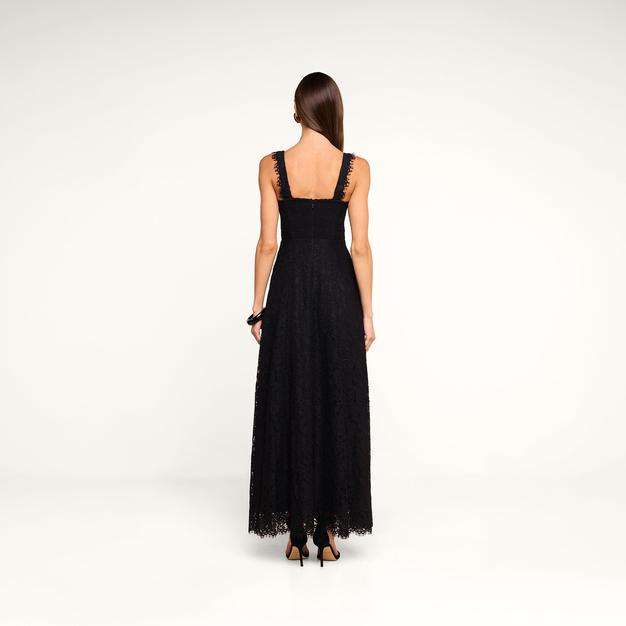 A woman in the Sutton Lace Maxi Dress, featuring black lace and a supportive fit, paired with black heels, stands facing away from the camera against a plain white background.
