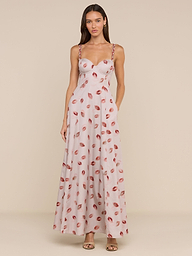 A woman wears the Sutton Maxi Dress, a light pink, floor-length gown with a fitted bodice, beaded straps, and a bold lipstick-kiss print. She pairs it with open-toe heels and her long brown hair is worn down.