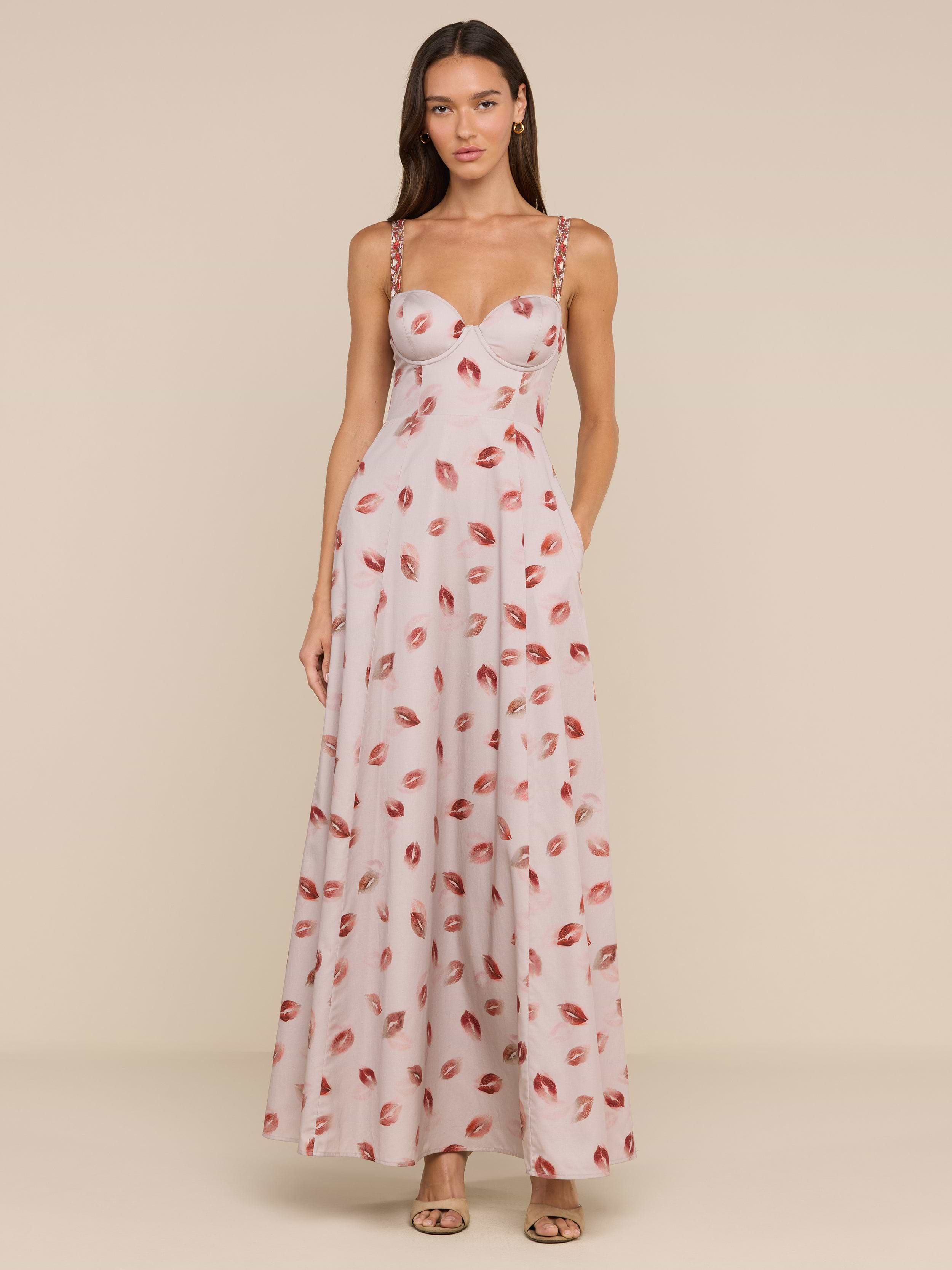 A woman wears the Sutton Maxi Dress, a light pink, floor-length gown with a fitted bodice, beaded straps, and a bold lipstick-kiss print. She pairs it with open-toe heels and her long brown hair is worn down.