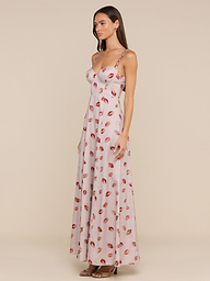 A woman stands against a plain beige background, wearing the Sutton Maxi Dress—a long, light pink gown featuring a lipstick-kiss print and delicate beaded straps.