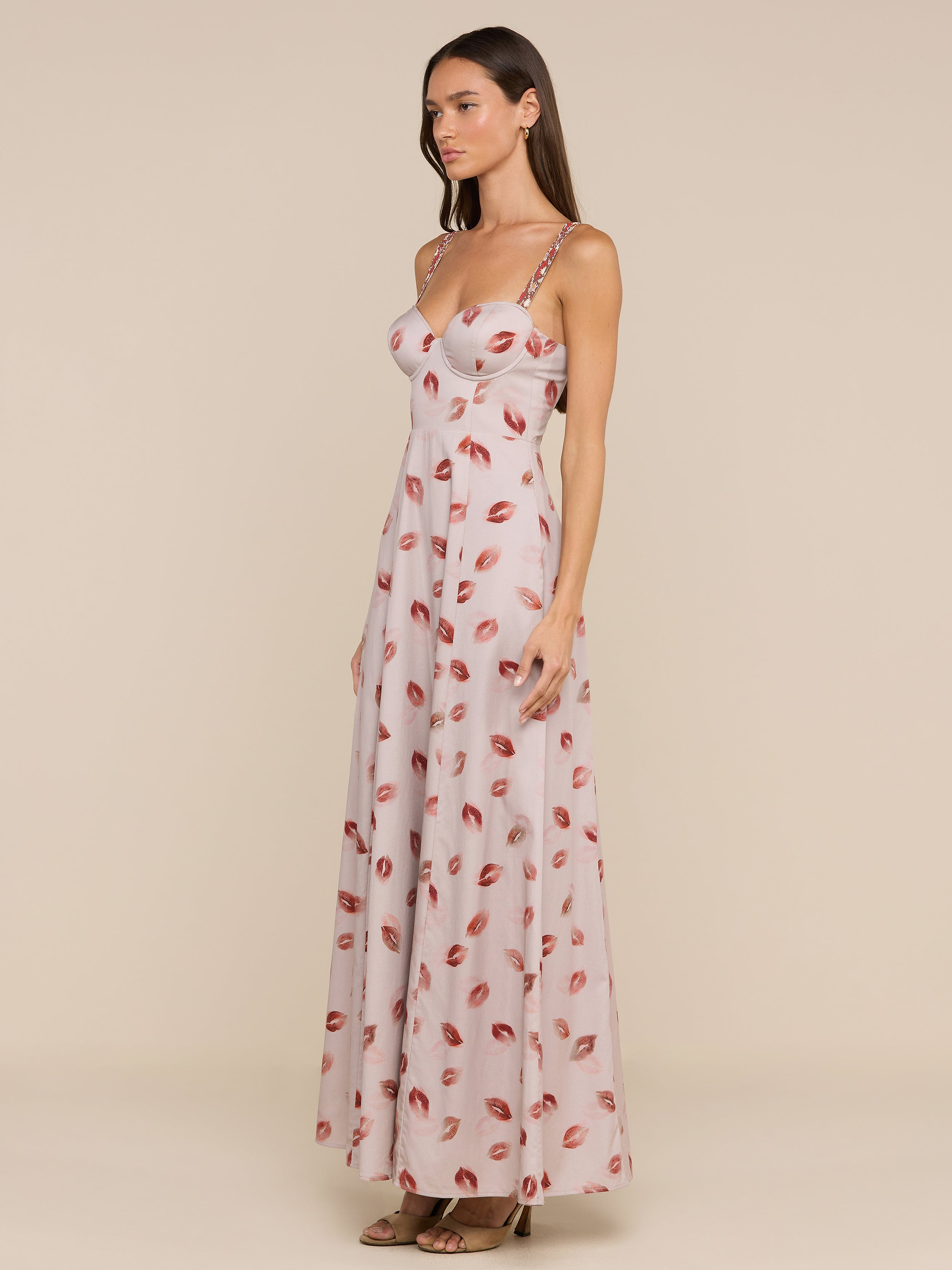 A woman stands against a plain beige background, wearing the Sutton Maxi Dress—a long, light pink gown featuring a lipstick-kiss print and delicate beaded straps.