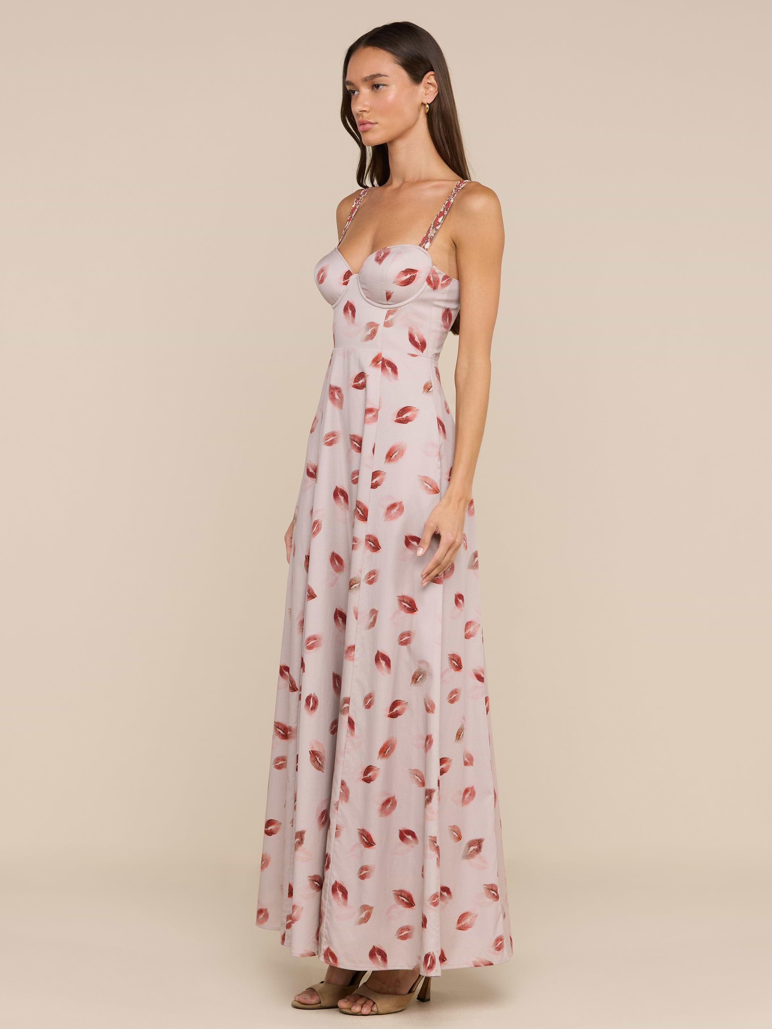 A woman stands against a plain beige background, wearing the Sutton Maxi Dress—a long, light pink gown featuring a lipstick-kiss print and delicate beaded straps.