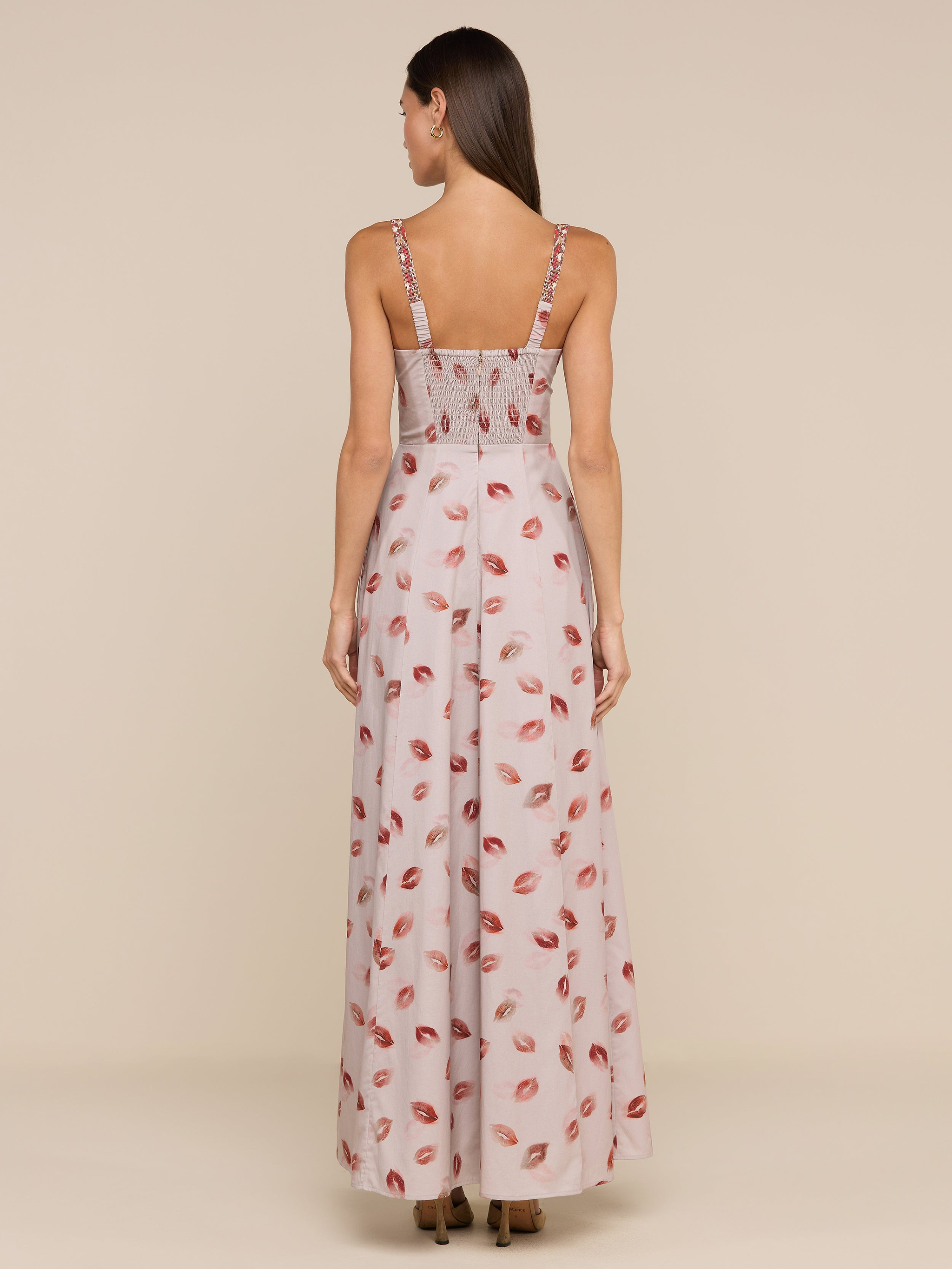 A woman with long brown hair, seen from behind, wears the Sutton Maxi Dress featuring a pink and red lipstick-kiss print and fitted bodice. The background is plain beige.