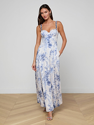 Una donna indossa il Sutton Maxi Dress senza maniche, un lungo abito bianco con motivi floreali blu e spalline di perline, in piedi su un pavimento di legno contro un muro bianco semplice. Lo abbina a sandali con tacco, orecchini e capelli sciolti.