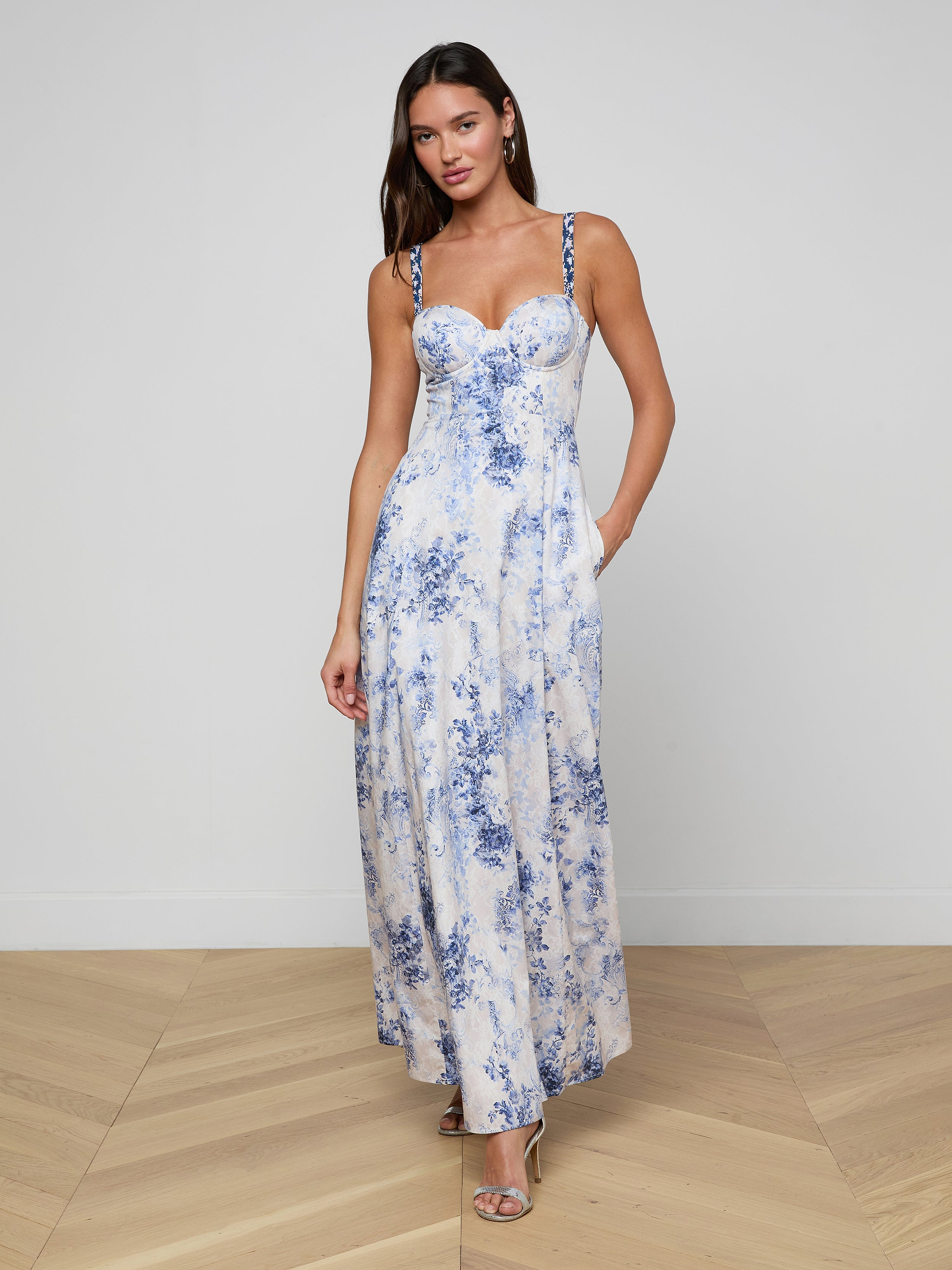Una donna indossa il Sutton Maxi Dress senza maniche, un lungo abito bianco con motivi floreali blu e spalline di perline, in piedi su un pavimento di legno contro un muro bianco semplice. Lo abbina a sandali con tacco, orecchini e capelli sciolti.
