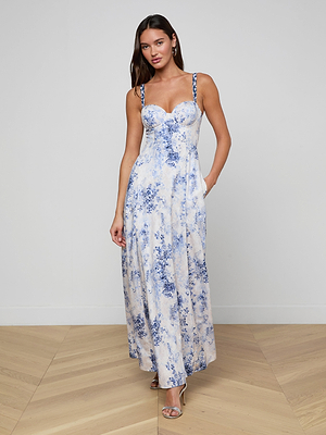 A woman wears the Sutton Beaded Strap Gown, a long, sleeveless spring-summer dress with blue floral patterns and thin beaded straps, paired with earrings and open-toe heels, standing on a wooden floor against a white wall.