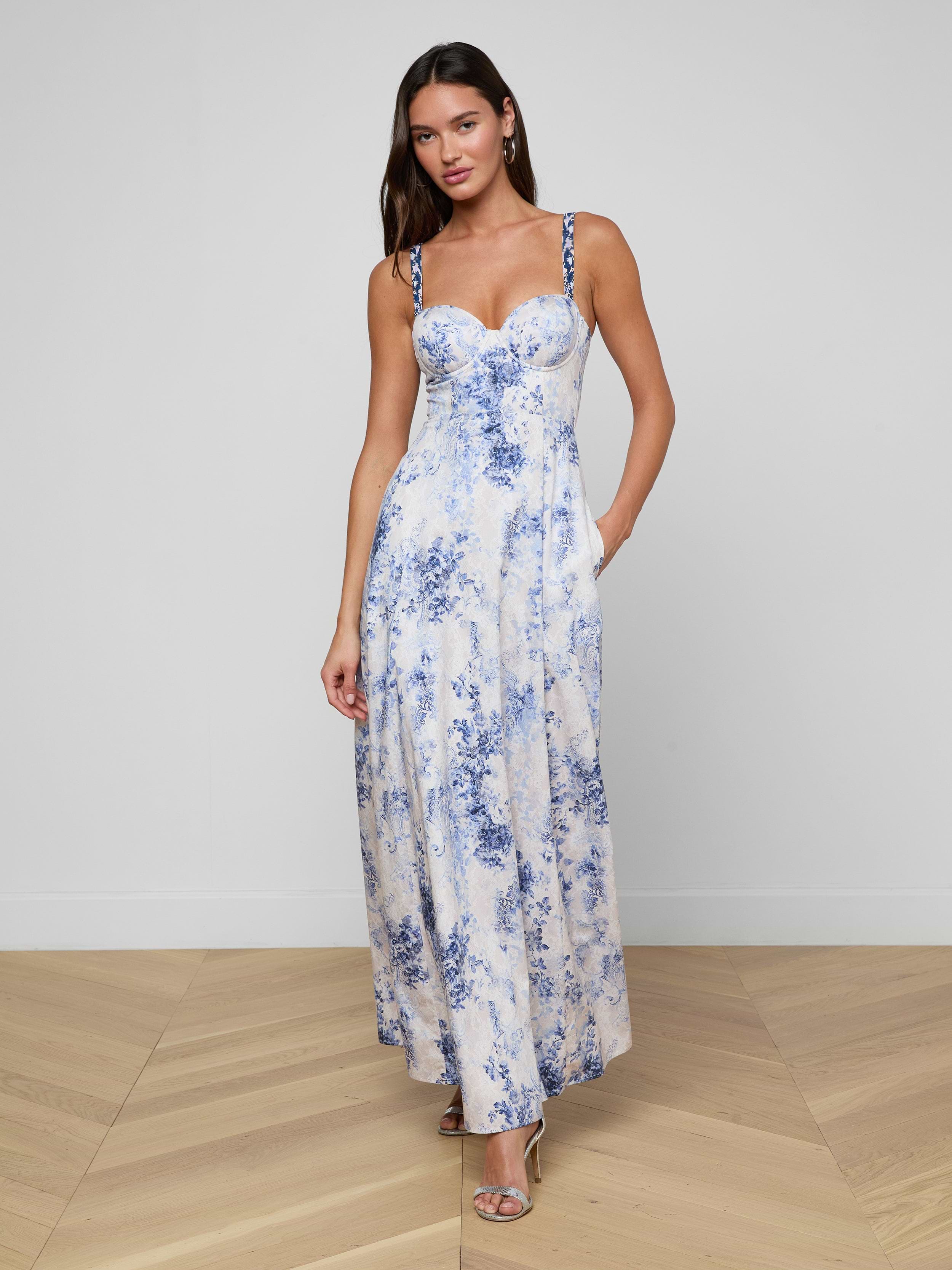 Una donna indossa il Sutton Maxi Dress senza maniche, un lungo abito bianco con motivi floreali blu e spalline di perline, in piedi su un pavimento di legno contro un muro bianco semplice. Lo abbina a sandali con tacco, orecchini e capelli sciolti.