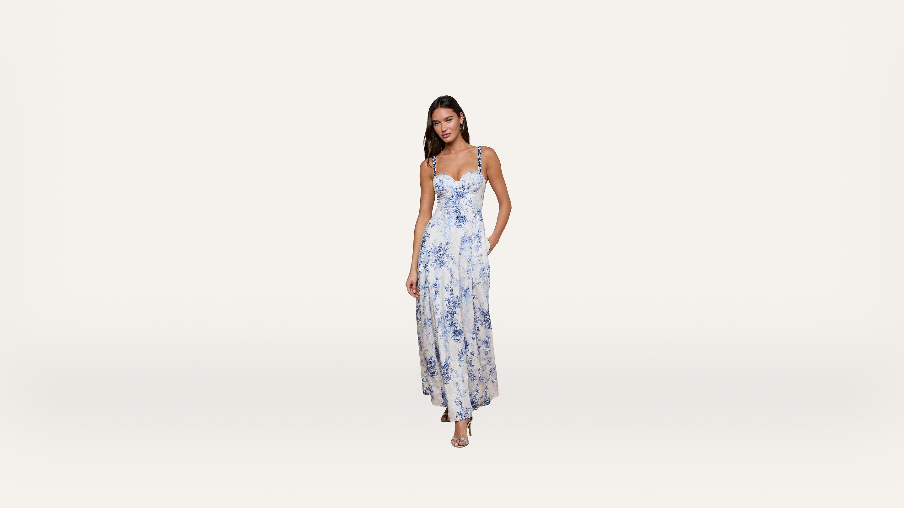 A woman stands wearing a long, sleeveless white dress with blue floral patterns and strappy sandals against a plain white background.