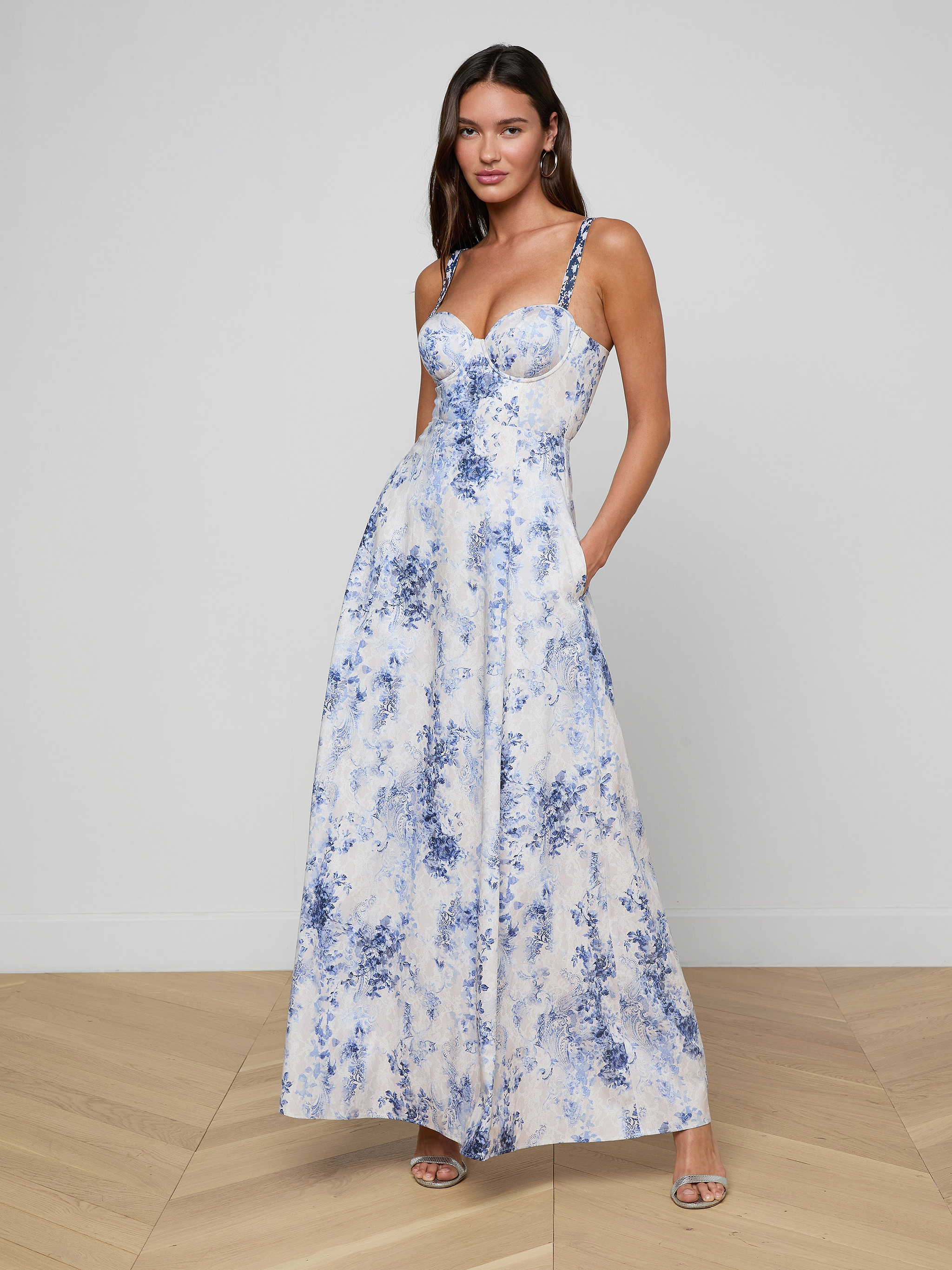 Una donna dai lunghi capelli castani si trova su un pavimento di legno chiaro e indossa il Sutton Maxi Dress, un maxi abito bianco senza maniche con stampe floreali blu. Ha le mani in tasca e lo abbina a dei tacchi argentati con una semplice parete bianca.