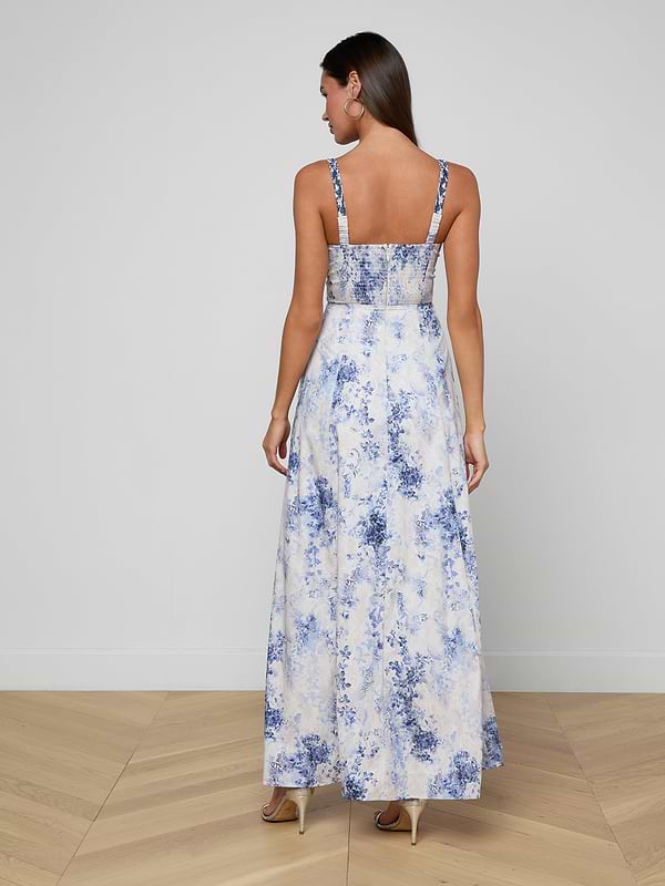Una donna con capelli lunghi, orecchini a cerchio e tacchi alti sta in piedi su un pavimento di legno chiaro contro un muro semplice, indossando il Sutton Maxi Dress senza maniche con motivi floreali blu e bianchi e delicate spalline di perline.