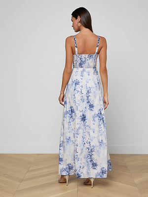 A woman stands on a wood floor facing away, wearing the Sutton Beaded Strap Gown—a long, sleeveless blue and white floral dress with thin straps and heels—against a plain light-colored wall.