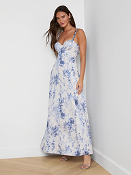 A woman with long brown hair stands against a white wall in the Sutton Maxi Dress, featuring blue floral patterns, beaded spaghetti straps, and open-toe heels. She poses casually.