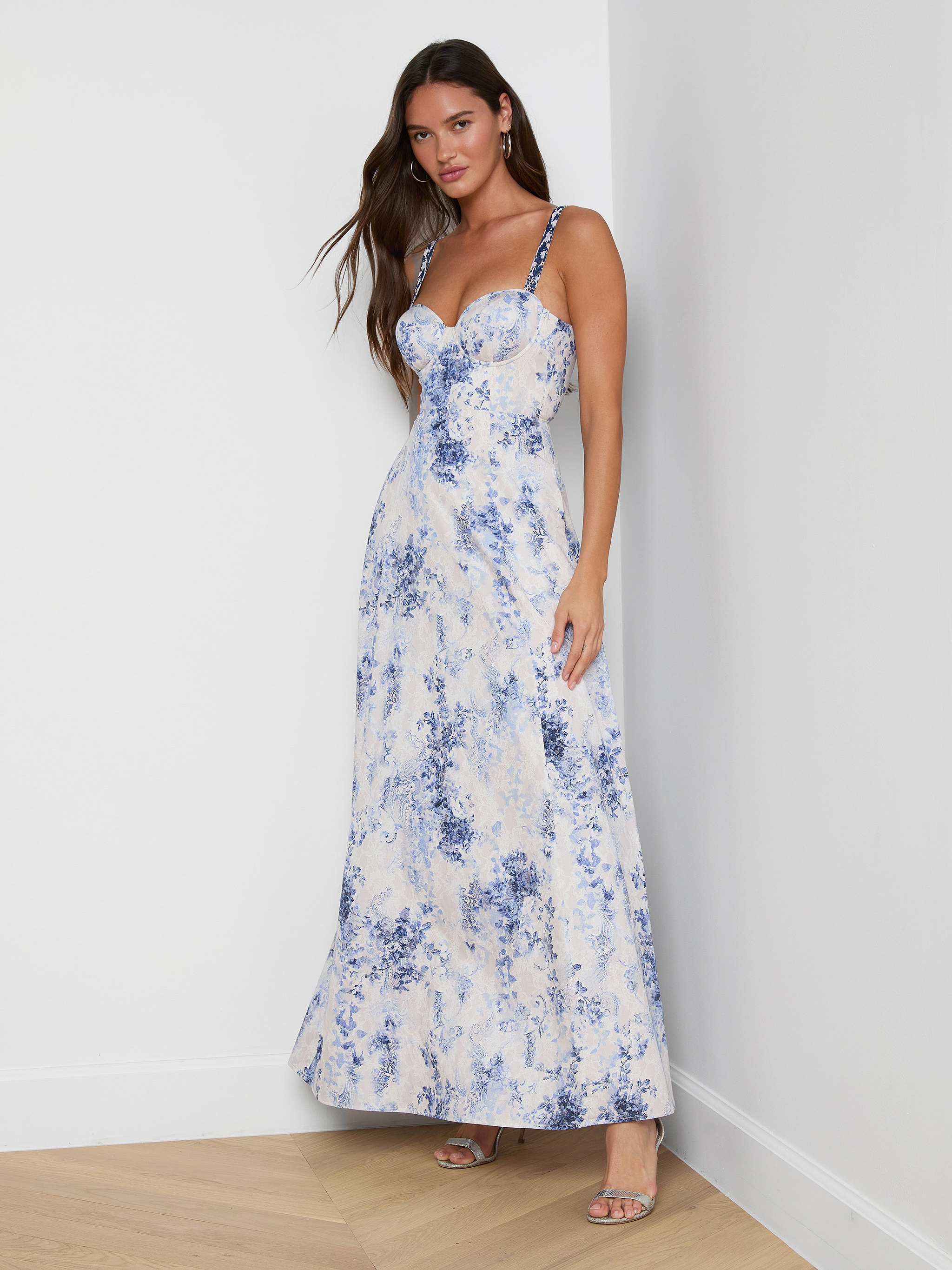 A woman with long brown hair stands against a white wall in the Sutton Maxi Dress, featuring blue floral patterns, beaded spaghetti straps, and open-toe heels. She poses casually.