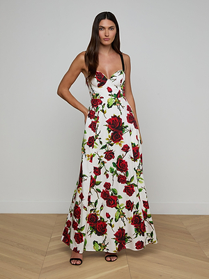 A woman with long brown hair poses indoors against a white wall, wearing the Sutton Beaded Strap Gown—a rose print maxi dress with black beaded adjustable straps—and black sandals, one hand casually placed in her pocket.
