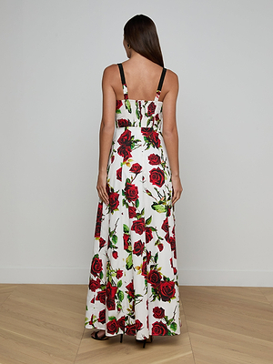 A woman with long brown hair stands on a wood floor facing away, wearing the Sutton Beaded Strap Gown—a white maxi dress with black beaded adjustable straps and a red rose print, set against a plain white wall.