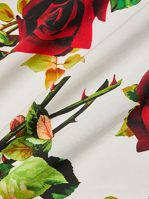 Close-up of the Sutton Beaded Strap Gown's white fabric, featuring a printed pattern of red roses with green leaves and stems. Gentle folds add texture, enhancing the floral design—ideal for this elegant rose print maxi dress.