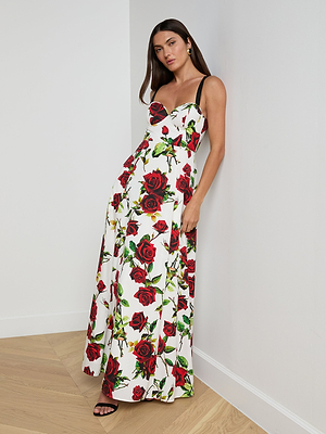 A woman stands in a corner wearing the Sutton Beaded Strap Gown, a long white maxi dress with red and green rose prints, beaded black straps, and a fitted bodice. She has long dark hair and is paired with black sandals.