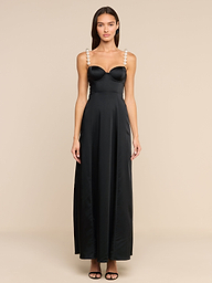 A woman stands against a plain white background, wearing the Sutton Maxi Dress—a long black satin evening gown with pearl-embellished straps—paired with black high-heeled sandals.