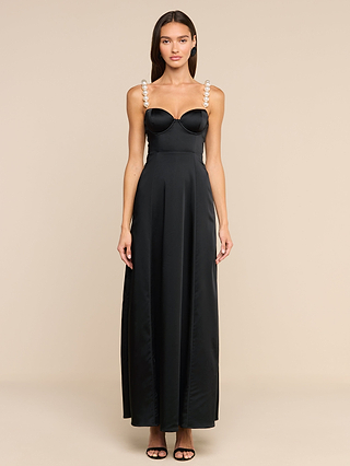 A woman stands against a beige background, wearing the Sutton Maxi Dress— a long black satin gown featuring simulated pearl straps— paired with black open-toe heels. Her hair is straight and down, and she faces forward with her arms relaxed by her sides.