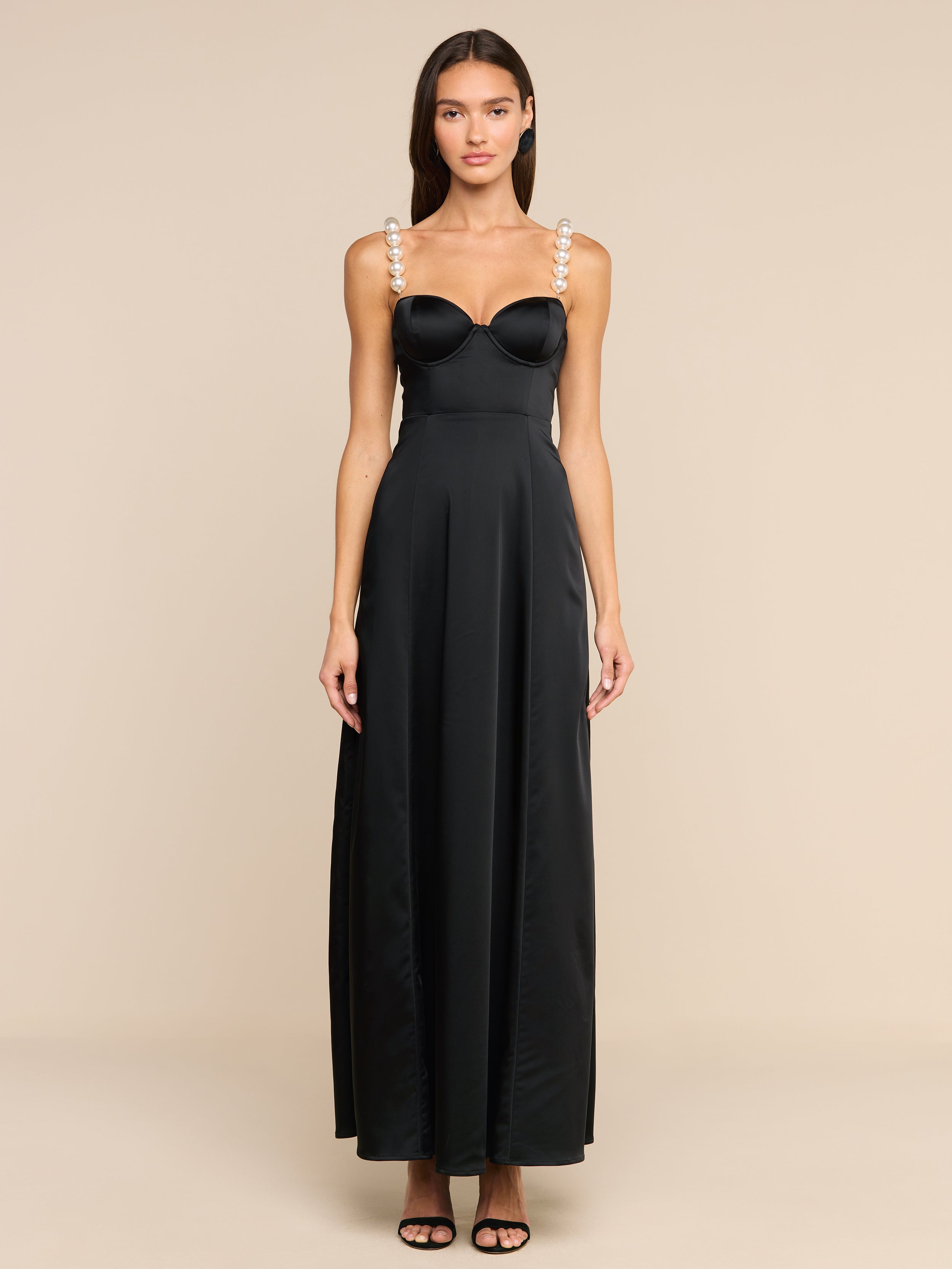 A woman stands against a plain white background, wearing the Sutton Maxi Dress—a long black satin evening gown with pearl-embellished straps—paired with black high-heeled sandals.