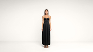 A woman stands against a plain white background, wearing a long black dress with spaghetti straps and black open-toe heels. She faces forward with her arms relaxed at her sides.