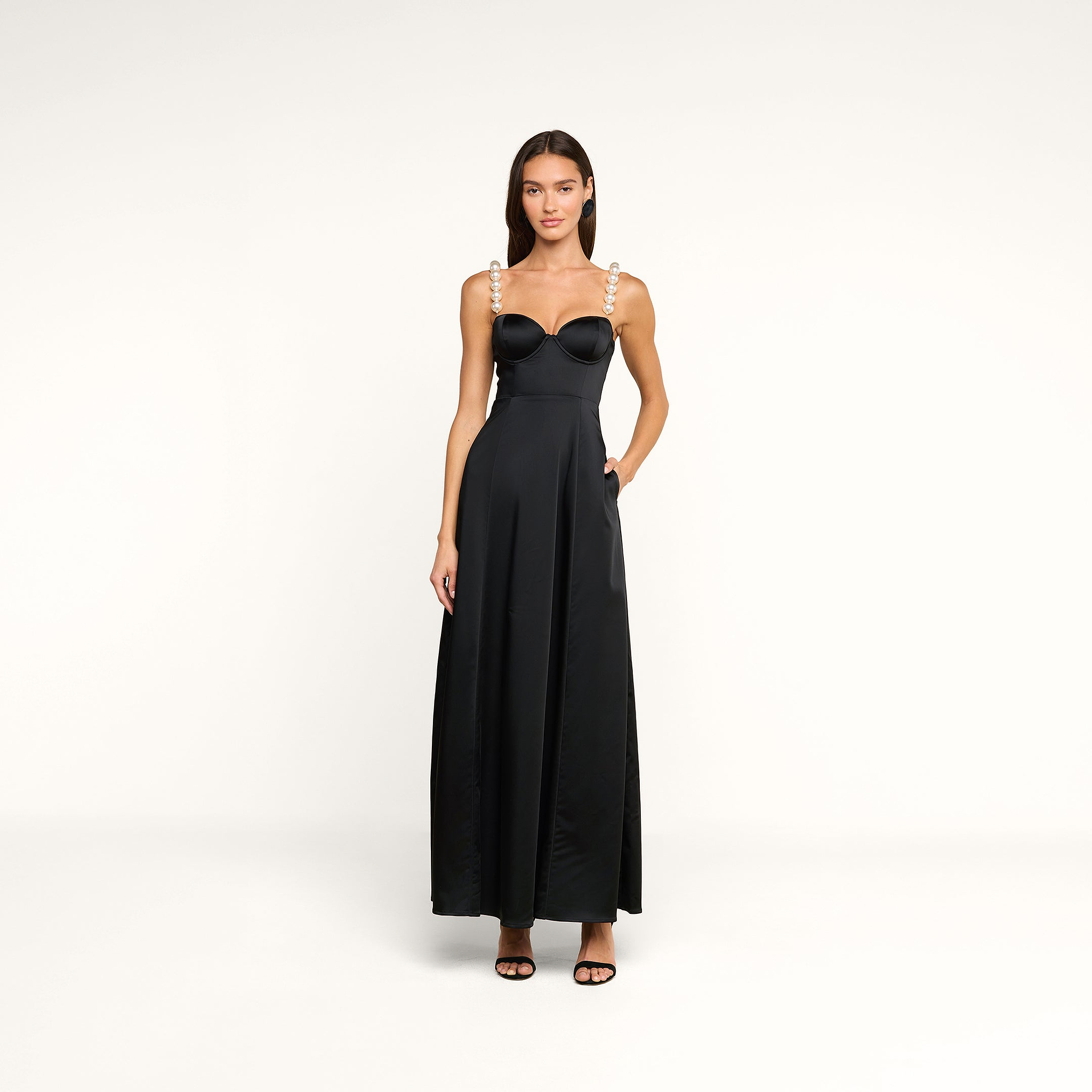 A woman stands facing forward in the Sutton Maxi Dress, a long black gown featuring pearl straps, paired with black heels and straight hair, set against a plain white background.